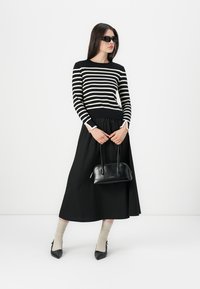 Femme en pull à rayures noires et blanches à manches longues, jupe midi noire, chaussettes beiges, escarpins noirs, tenant un sac à main noir, portant des lunettes de soleil noires.