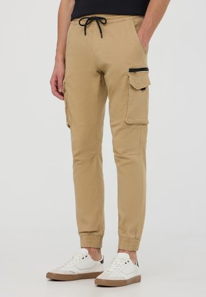 Pantaloni cargo khaki con vita elasticizzata e cordino, dotati di tasche con zip e tasche a patta, gambe affusolate e polsini a coste, indossati con sneakers bianche.