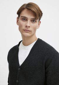 Svart strikket cardigan med V-halsdesign, som har tre svarte knapper og en teksturert overflate, lagdelt over en hvit T-skjorte.
