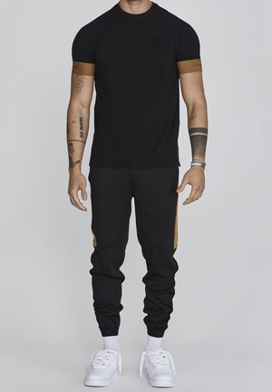 Mann trägt schwarzes Kurzarmhemd mit braunen Bündchen, schwarze Jogginghose mit braunen Seitenstreifen, weiße Socken und weiße Turnschuhe, steht aufrecht.