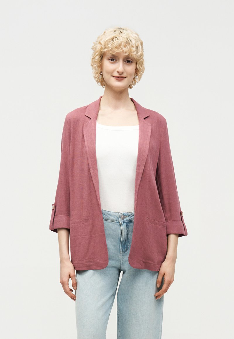 Άτομο με ξανθά σγουρά μαλλιά που φοράει ροζ σακάκι με ανοιχτό μπροστινό μέρος, λευκό τοπ και ανοιχτόχρωμο μπλε τζιν, στέκεται μπροστά σε απλό φόντο.