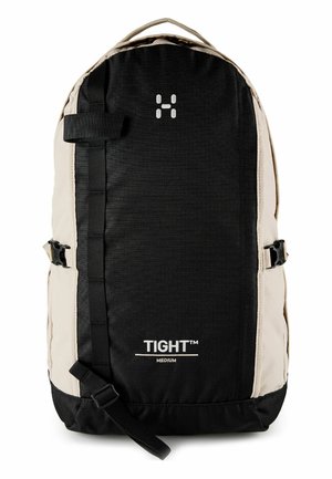 Zaino di media dimensione nero e beige con cinturini anteriori, fibbie laterali, maniglia superiore e la scritta "TIGHT™ MEDIUM" stampata sul pannello frontale.