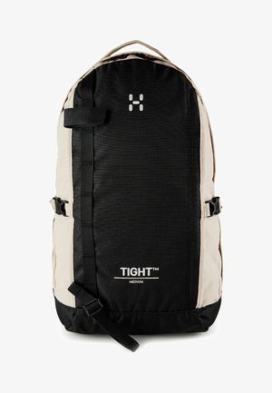 Sac à dos de taille moyenne noir et beige avec des sangles à l'avant, des boucles latérales, une poignée supérieure, et "TIGHT™ MEDIUM" imprimé sur le panneau avant.