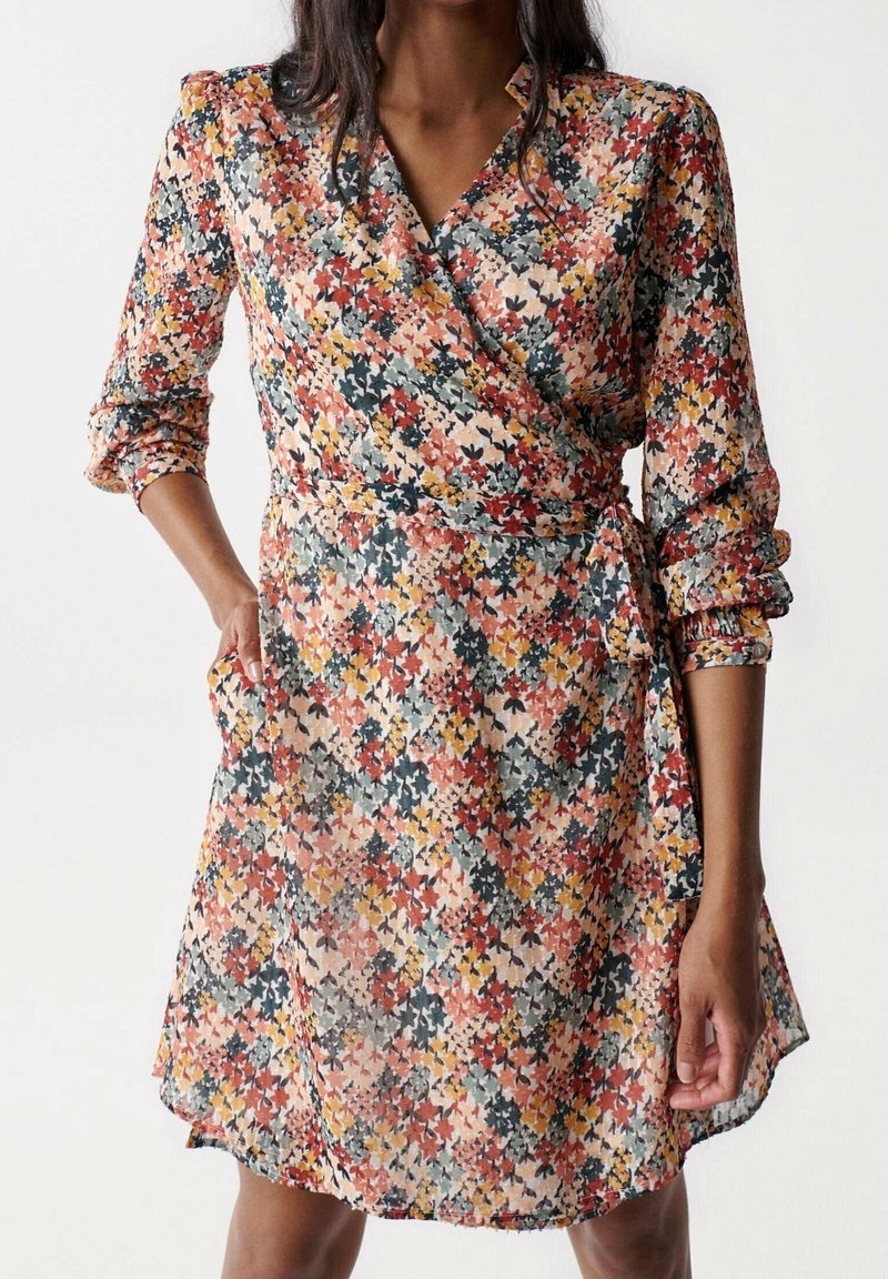 Femme portant une robe portefeuille mi-longue à manches longues avec un motif floral multicolore en rouge, jaune, gris et noir.