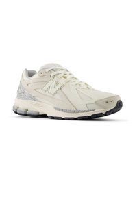 Baskets de sport blanc cassé avec tige en mesh et synthétique, semelle coussinée et semelle extérieure noire, avec un détail de logo "N" sur le côté.