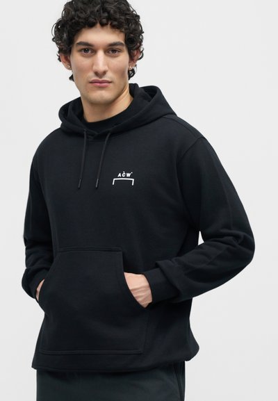 Άνδρας με σγουρά μαλλιά που φοράει μαύρο φούτερ με λευκό λογότυπο ACW, τα χέρια στην μπροστινή τσέπη, κοιτάζει μπροστά σε απλό φόντο.