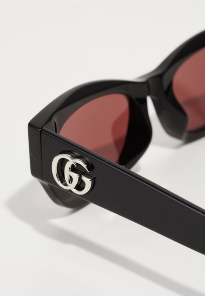 Las gafas de sol negras cuentan con lentes rojos, un diseño angular y un acabado brillante. Destaca el acento plateado del logo GG en la patilla.