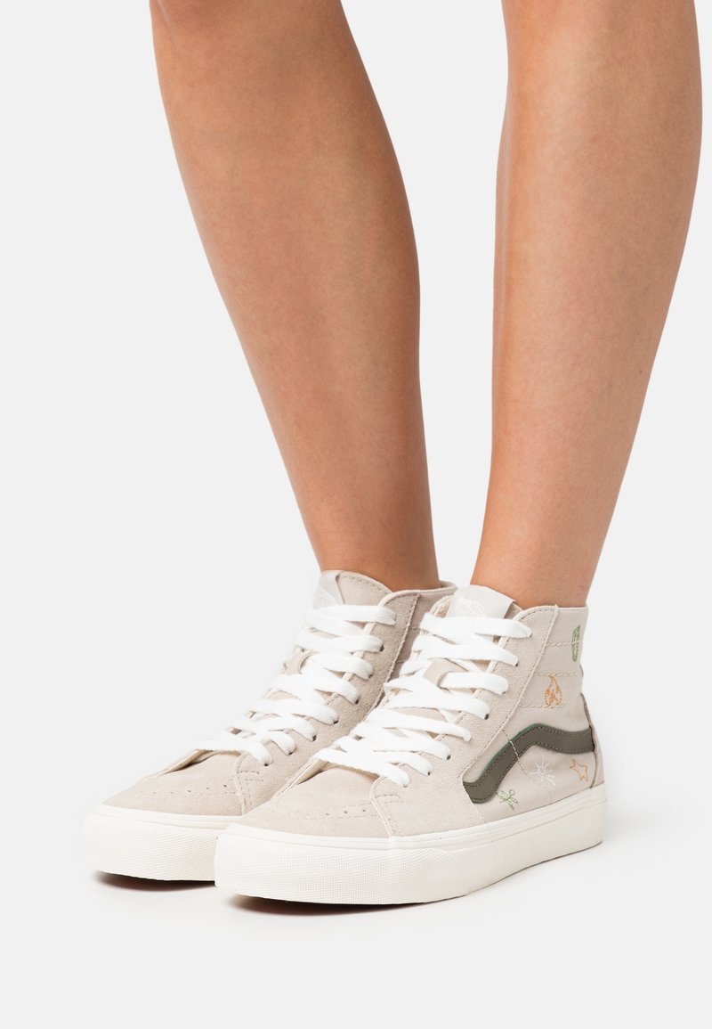 Baskets montantes beige clair avec lacets blancs, semelles blanches, bande latérale foncée et petits motifs brodés de feuilles et de plantes portées sur jambes nues.