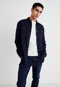 Chemise en denim foncé avec une fermeture à boutons sur le devant, deux poches poitrine et des coutures oranges contrastantes. Portée par-dessus un t-shirt blanc et assortie à un pantalon en denim.