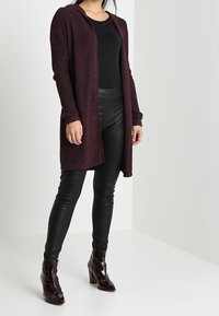 Cardigan en maille bordeaux à ouverture sur le devant, manches longues et texture côtelée, associé à des leggings en cuir noirs et des bottines vernies.