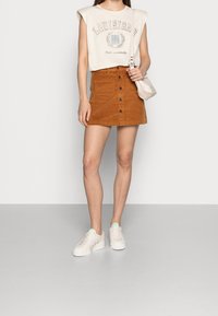 T-shirt cropped beige clair avec l'inscription "Louisiana", associé à une mini-jupe en velours côtelé couleur rouille avec des boutons à l'avant, porté avec des baskets blanches.