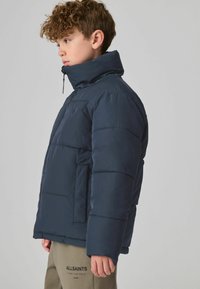 Jūras spēku pūkains apģērbs ar augstu apkakli, ar rāvējslēdzēju un sānu kabatām. Iespējas ietver quilted dizainu un gludu tekstūru, uzvilkts virs bēšām biksēm.