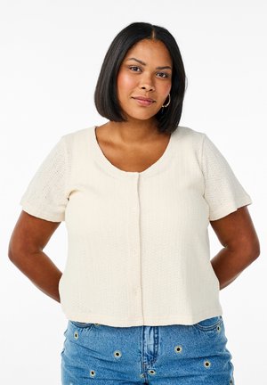 Light cream short-sleeve top with a textured pattern, round neckline, and front buttons. Paired with blue jeans featuring decorative accents.
