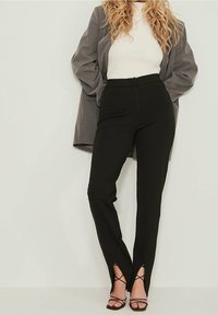 Mujer con cabello rubio rizado que lleva un blazer gris, una blusa blanca de canalé, pantalones negros de tiro alto con aberturas frontales y tacones negros con tiras.
