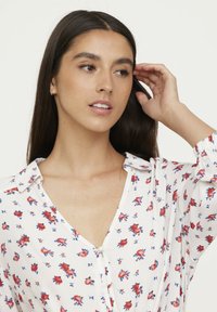 Weißes Bluse mit kleinen roten und blauen Blumenmustern, Knopfleiste und einem Kragen. Strukturierter Stoff mit leichten Falten an den Schultern.