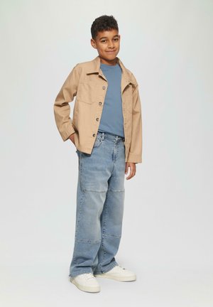 Lichtbruin button-up jack over een blauwe T-shirt, gecombineerd met losse lichtblauwe jeans en witte sneakers. Soepele texturen, casual pasvorm.
