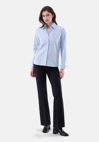 Camicia azzurro chiaro con colletto a punta, indossata con pantaloni neri slim-fit e stivali alla caviglia con tacco, su uno sfondo bianco semplice.