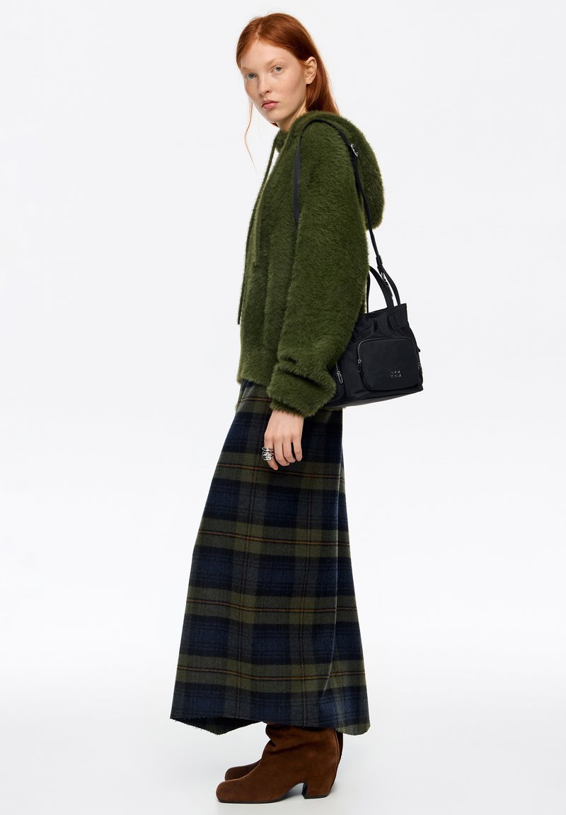 Zielona, puszysta kurtka z kapturem, długa spódnica w kratę w ciemnoniebieskim i zielonym kolorze, brązowe zamszowe botki na grubym obcasie oraz czarna torebka z zamkami błyskawicznymi.