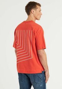 Maglietta rossa a maniche corte con un motivo geometrico bianco sul retro, scollatura a girocollo e vestibilità ampia, abbinata a jeans blu.