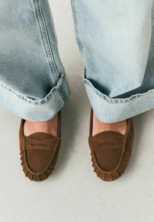 Light blue cuffed jeans worn with brown suede moccasin loafers on a light gray floor.
