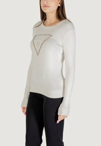 Suéter crema con un cuello y puños acanalados. Presenta un logo triangular con strass en la parte frontal. Combinado con pantalones negros.