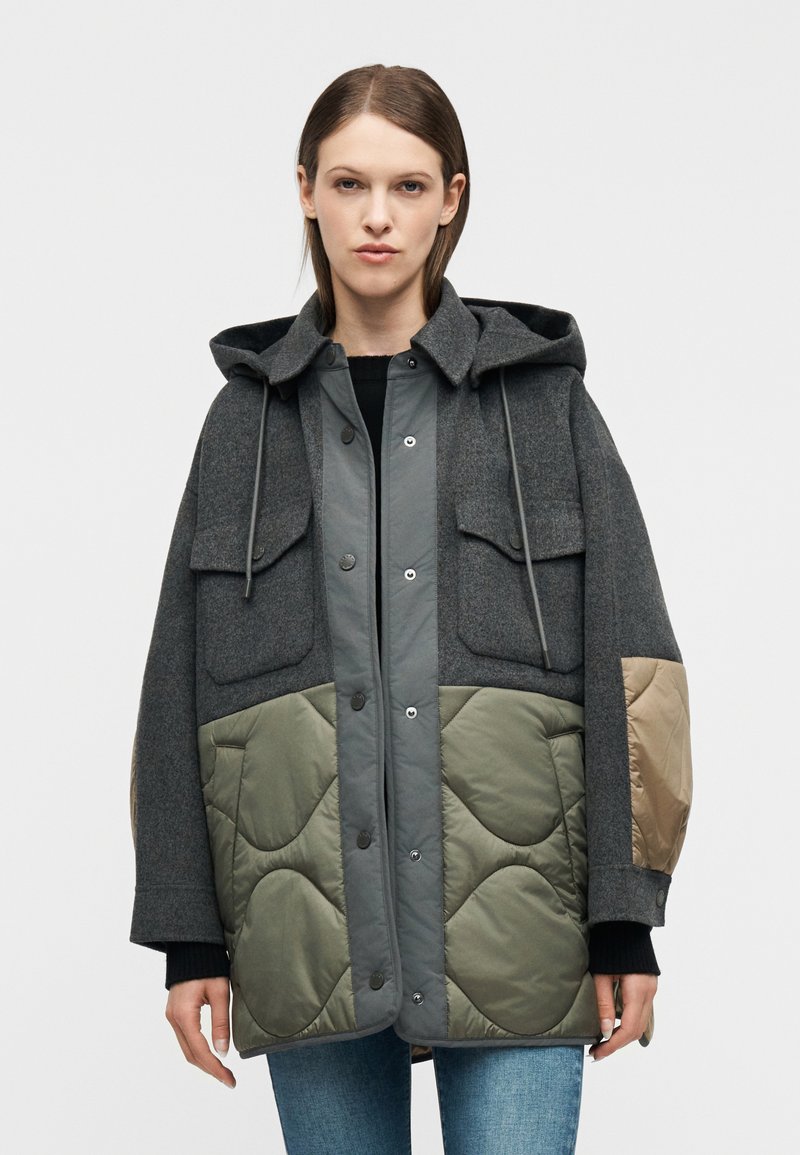 Γυναίκα που φοράει μια φαρδιά ζακέ τα με κουκούλα, με γκρι μάλλινο πάνω μέρος, καπιτονέ χακί κάτω τμήμα και ταμπά επιθέματα στους αγκώνες, που στέκεται μπροστά σε λευκό φόντο.