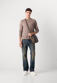 Pulover tricotat de culoare maro deschis, blugi albaștri și adidași albi. O mică geantă crossbody de culoare maro cu o curea este ținută în mana stângă.