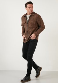 Giacca in misto lana marrone con chiusura a zip, due tasche sul petto e colletto classico, abbinata a pantaloni neri slim-fit e stivali neri con lacci.