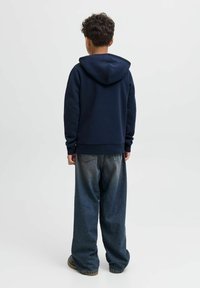 Person mit kurzen lockigen Haaren, die einen dunkelblauen Hoodie, weit geschnittene ausgebleichte Jeans und braune Schuhe trägt, steht mit dem Rücken zu einem hellen Hintergrund.