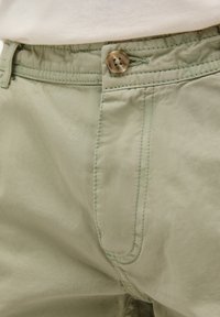 Pantaloni scurți din bumbac verde deschis, cu închidere cu nasture, buzunare laterale și cusături contrastante pe margini. Textură moale și design casual.