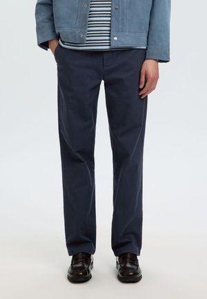 Homme portant un pantalon droit bleu marine foncé, des chaussures marron foncé, une chemise rayée et une veste bleue, debout avec une main dans la poche.