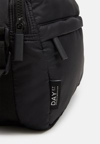 Borsa nera a sacco realizzata in nylon con zip e tracolla nera. Presenta un piccolo etichetta con testo bianco che dice "DAY ET". Superficie semplice e texturizzata.