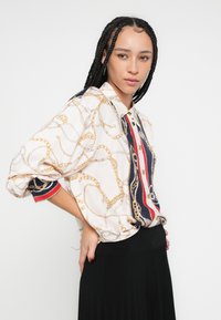 Bijela košulja s uzorkom zlatnog lanca, mornarskoplave i crvene pruge na ovratniku i manžetama. Mekani materijal, opuštenog kroja, s kopčanjem na gumbe.