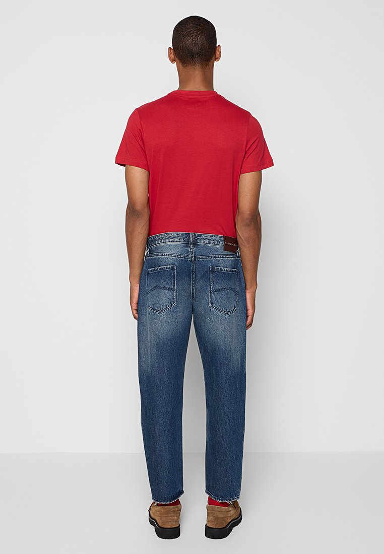 Mężczyzna ubrany w czerwony krótki rękaw, niebieskie dżinsy i brązowe buty, stojący tyłem na jednolitym jasnoszarym tle.