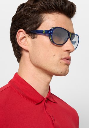 Uomo che indossa una camicia rossa con colletto e occhiali da sole ovali blu con accenti verdi, rivolto verso destra su uno sfondo semplice.