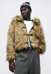 Casaco curto de pele sintética em castanho claro com acabamento texturizado, apresenta ferragens prateadas, fechos de mola e uma silhueta ajustada. Combinado com calças de risca fina.