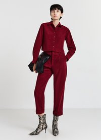Weinrotes Cordhemd mit Knöpfen, passende schmal zulaufende Hose und spitze Stiefeletten mit Schlangenmuster. Hält eine schwarze Clutch-Tasche.