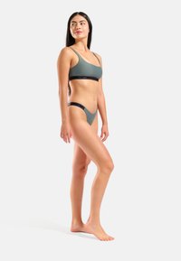 Conjunto de bikini verde esmeralda con una textura suave y mate. El top tiene tirantes delgados y una banda negra con logo; la parte inferior es de corte minimalista.