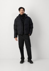Doudoune noire avec un col haut, fermeture éclair et design matelassé ; portée sur un col roulé noir et associée à un pantalon cargo noir et des chaussures.