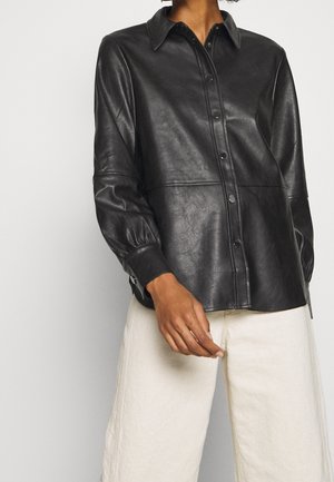 Personne portant une chemise à boutons en similicuir noir avec manches longues et col, associée à un pantalon large blanc cassé.