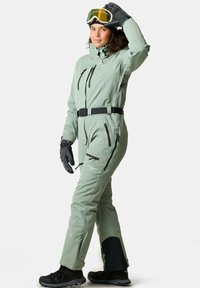 Kombinezon narciarski w jasnym zielonym kolorze, z zamkiem błyskawicznym z przodu, wieloma kieszeniami, paskiem i czarnymi akcentami. Zawiera rękawice i gogle.