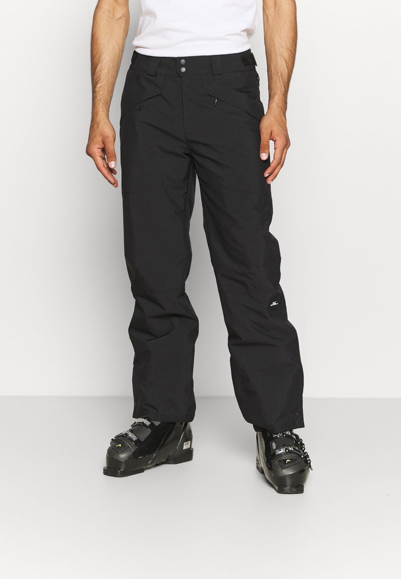 Homme portant un pantalon de ski noir et des chaussures de ski noires, debout devant un fond clair uni.