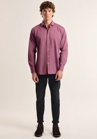 Camisa de manga larga y botonadura en un tejido púrpura suave, con un corte ajustado. Combinada con jeans negros ajustados y zapatos de ante marrón.