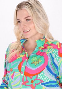 Camicia multicolore con colletto arricciato, chiusura a bottoni e vivaci motivi floreali in tonalità di rosa, verde e blu.