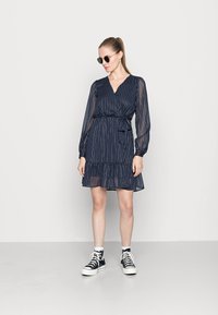 Vestido azul-marinho com mangas longas transparentes, decote em V e cintura cingida. Apresenta uma barra com babados e riscas verticais subtis. Combinado com sapatilhas pretas.