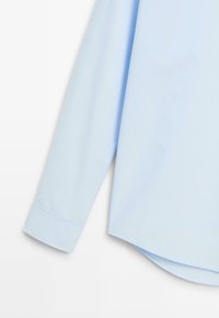 Hellblaues langärmliges Hemd mit geschwungenem Saum auf weißem Hintergrund, zeigt Manschettendetail und glatte Stoffstruktur.