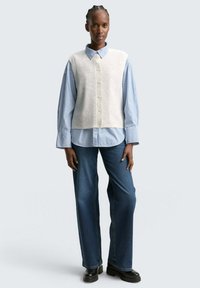 Gilet en maille gris clair avec des boutons sur une chemise à rayures bleues et blanches, associé à un jean large bleu foncé et des bottes à plateforme noires.