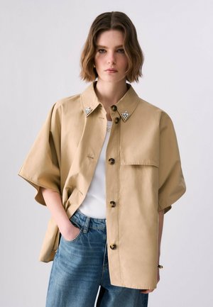 Mujer joven con chaqueta beige de manga corta abotonada con detalles de perlas en el cuello, camiseta blanca y vaqueros azules, manos en los bolsillos, fondo neutro.