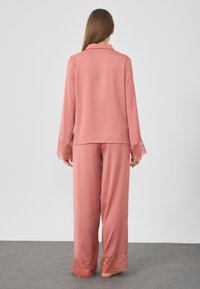 Svetloružový pyžamový set obsahuje dlhé rukávy s čipkovými akcentmi a zodpovedajúce nohavice. Mäkká, textúrovaná tkanina s voľným strihom.