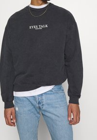 Felpa grigia scura con polsini e orlo a costine, con la scritta "EYES TALK" e "KAOTIKO" stampata in bianco sulla parte anteriore. Indossata sopra una camicia bianca.